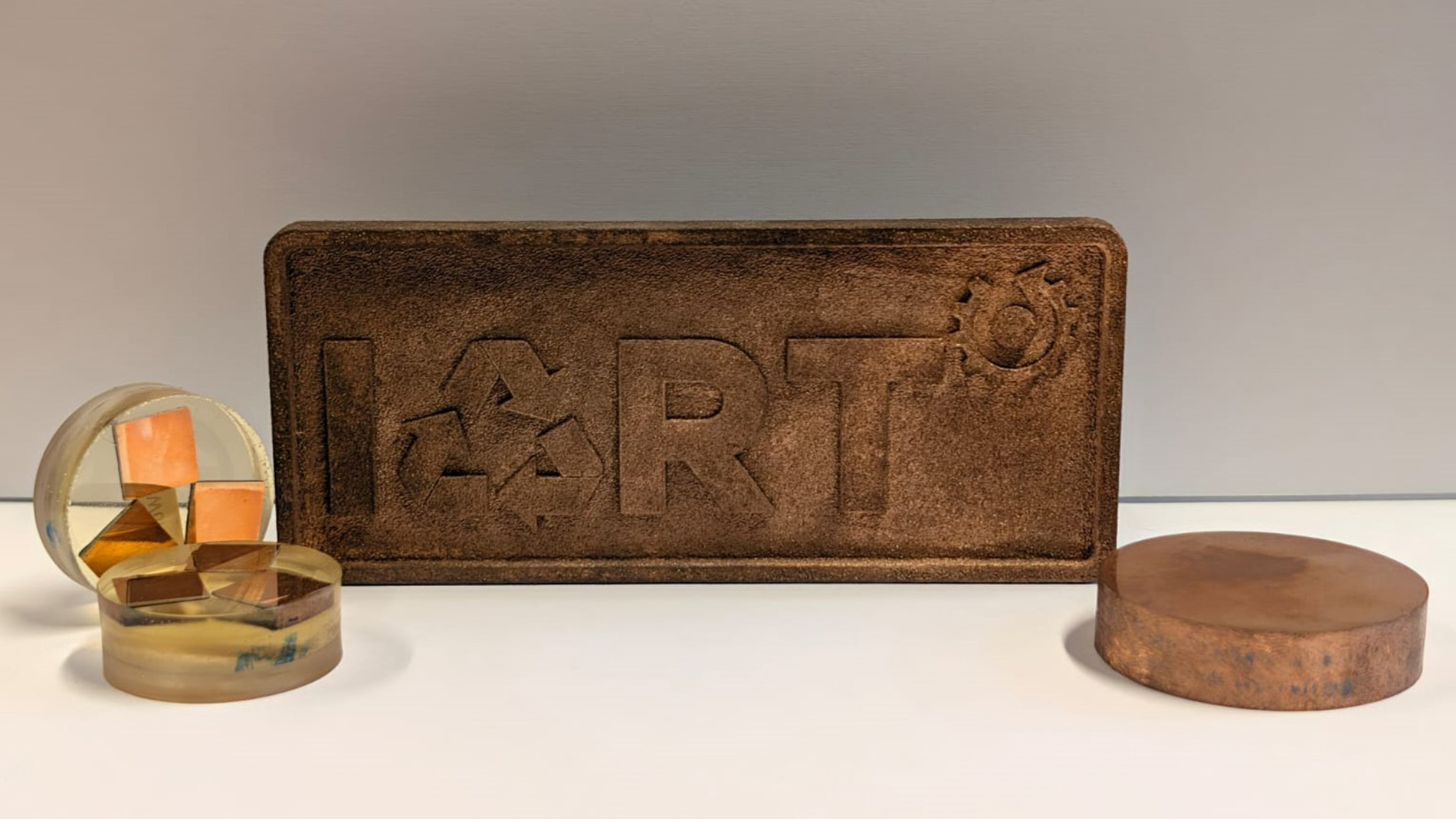Gedrucktes IART-Logoschild, zwei in Kunstharz eingebettete Würfelproben sowie eine Probe aus Sinterversuchen, gefertigt aus recyceltem Kupferpulver.